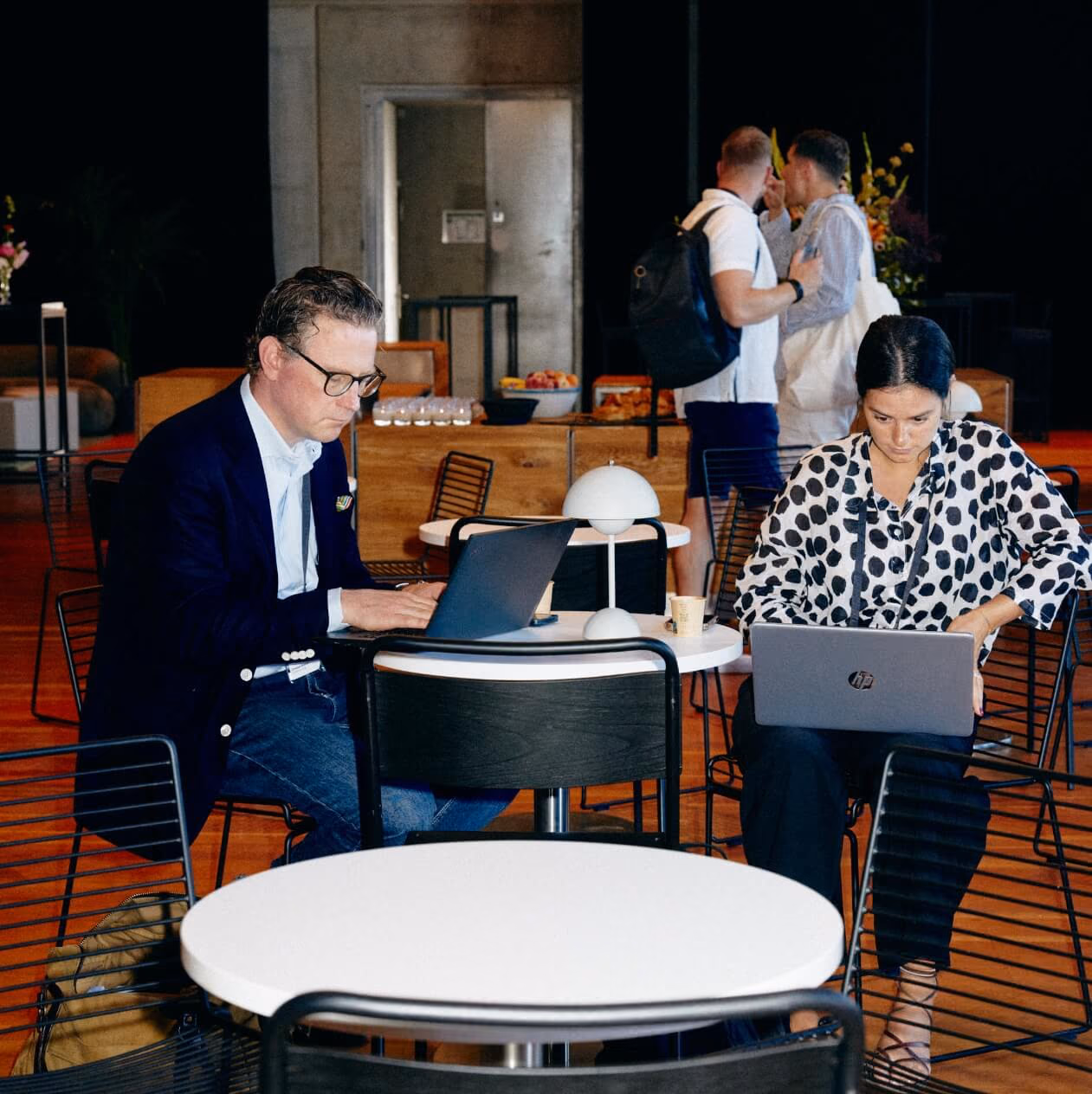 Zwei Personen arbeiten an runden Cafétischen an Laptops, während sich andere dahinter an einer Snacktheke unterhalten.