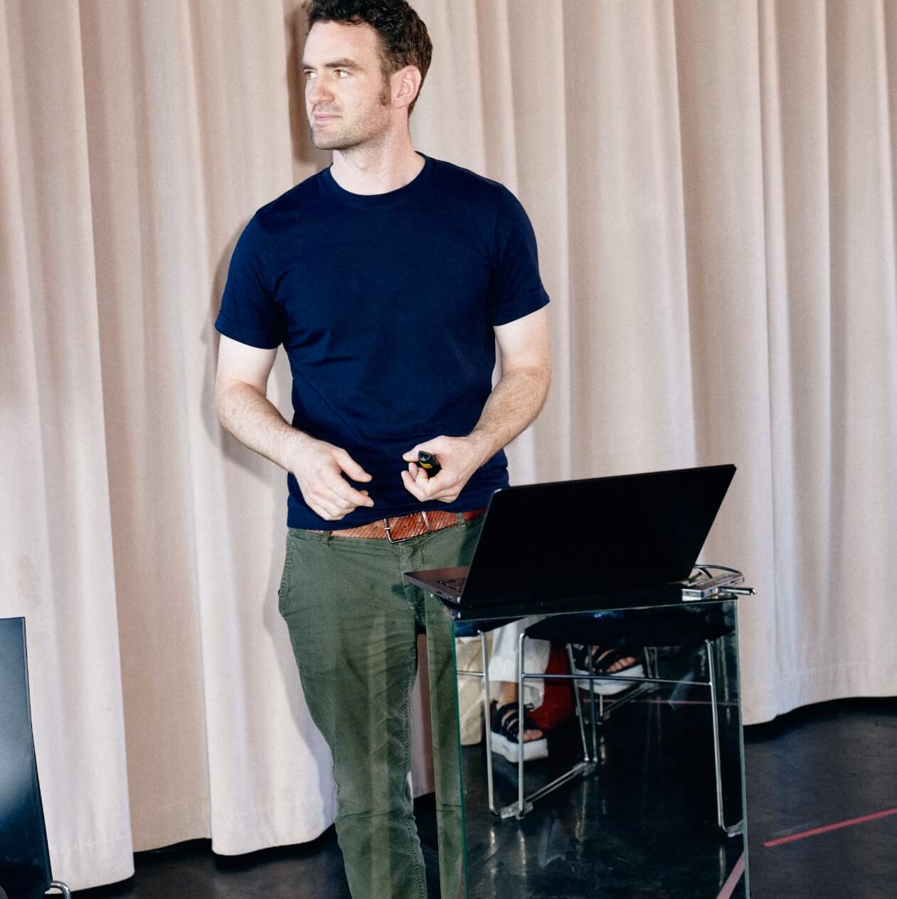 Ein Sprecher im dunklen T-Shirt hält einen Presenter neben einem Laptop an einem Glaspult.