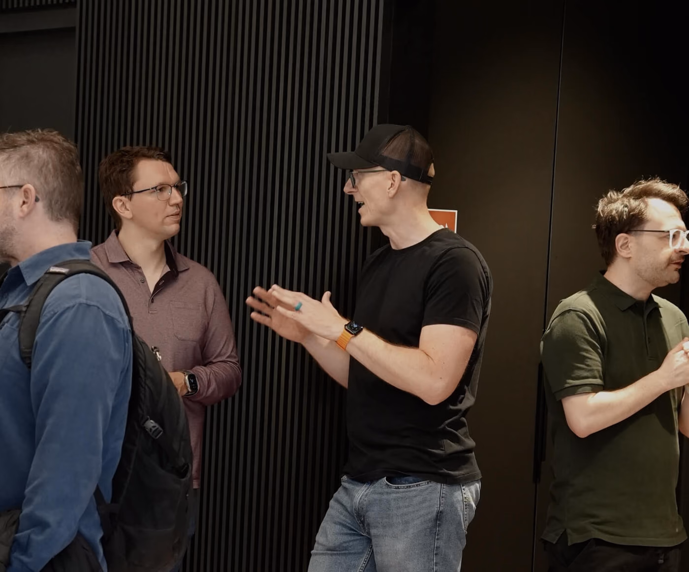 Eine kleine Gruppe unterhält sich in einem dunklen Raum, eine Person mit Cap gestikuliert mit beiden Händen, während die anderen zuhören.