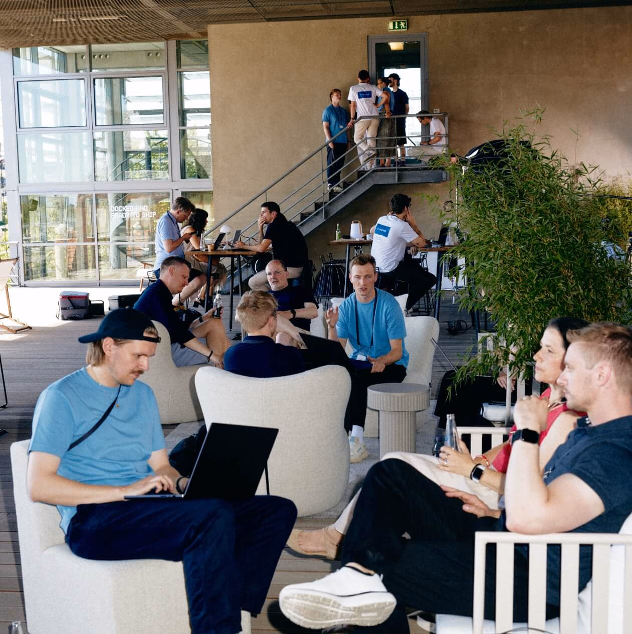 Menschen unterhalten sich und arbeiten an Laptops in Loungesesseln auf einer überdachten Terrasse, dahinter weitere Gäste auf einer Treppe.