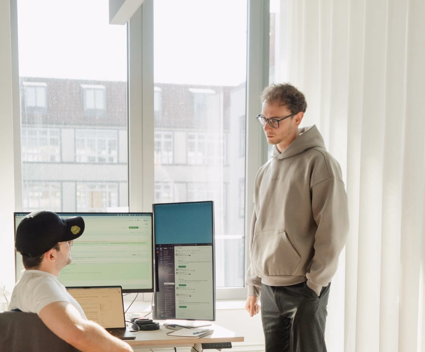 Eine Person im Hoodie steht an einem hellen Fenster und schaut einem Kollegen zu, der an einem Schreibtisch mit mehreren Bildschirmen arbeitet.