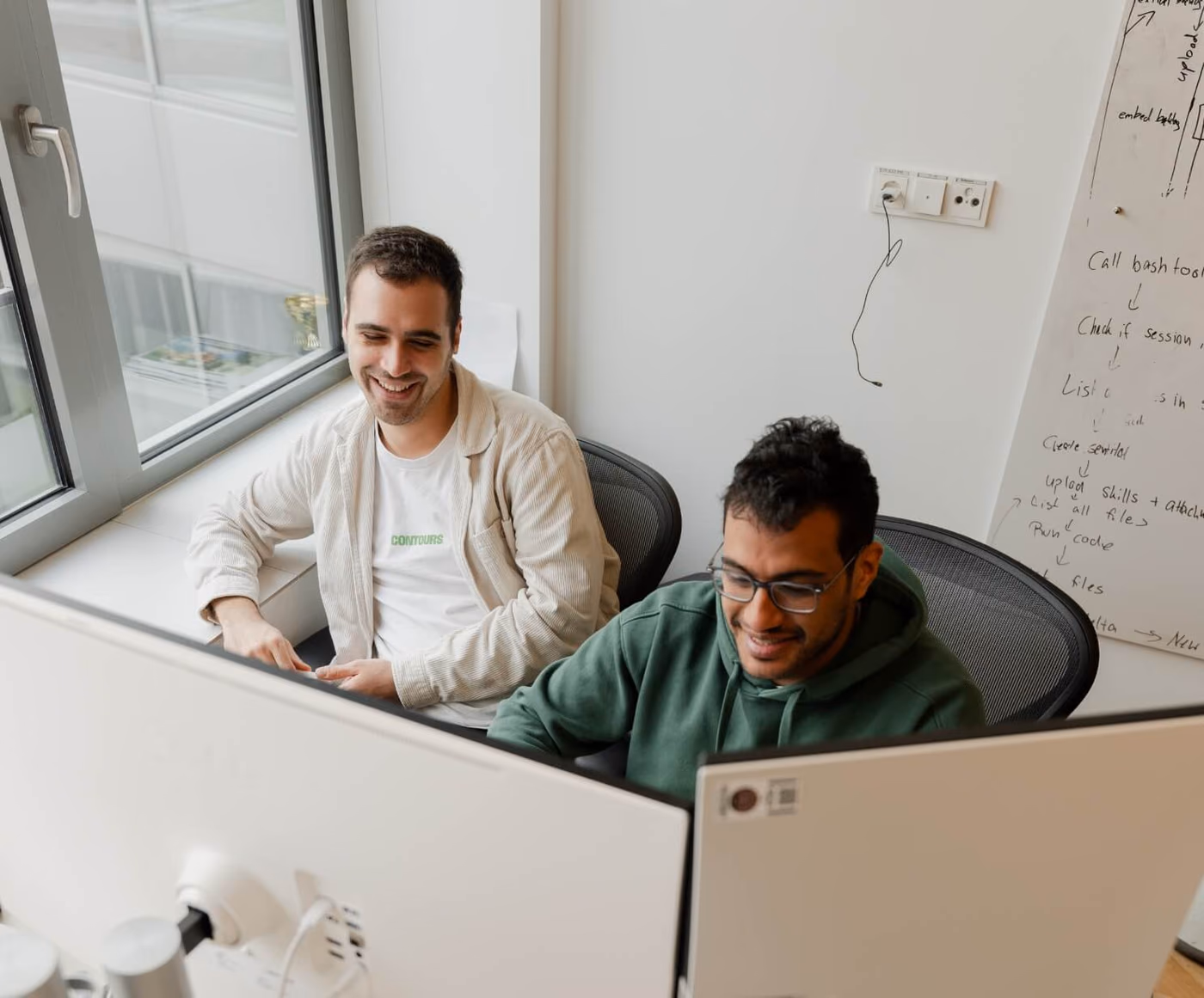 Zwei Personen lächeln an einem gemeinsamen Schreibtisch auf ihre Bildschirme, hinter ihnen ein handgezeichneter Workflow auf einem Whiteboard.